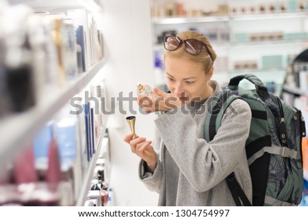Blond young female traveler wearing coat and travel backpack choosing perfume in airport duty free store. Casual lady testing and buying cosmetics on the go in a beauty store.