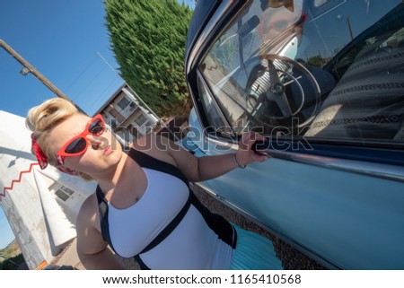 Blonde adult female with a 1950s vintage pin up hairstyle stands near an abandoned vintage car. Girl wearing red cateye vintage sunglasses. 