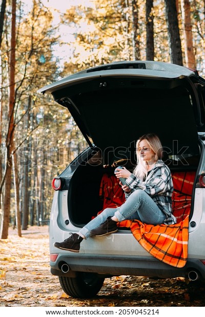 a blonde\
adult girl in a plaid shirt is drinking coffee sitting in the open\
trunk of a car. picnic in the\
car.