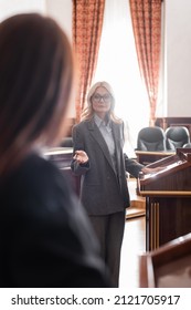 Blonde Advocate Pointing With Hand While Questioning Blurred Witness In Court