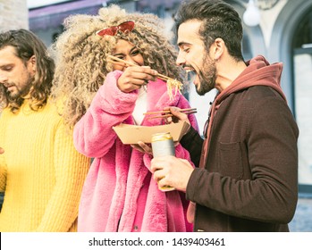 A Blonde Afro American Woman Take Noodles In The Mouth Of Her Friends Standing In The Streets. Young People Having Time Together Eating Asian Food Together. Leisure Activities Lifestyle Concept.