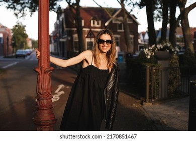 Blonde Appearance Woman With Two Thin Braids Holds Pillar In Fresh Air With Her Hand. Girl Smiles Cutely, Circling Around Post, Dressed In Black Dress.