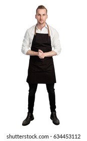 Blonde Attractive Chef Wearing A White Shirt And Black Apron Standing With His Hands Clasped Against A White Background Looking At Camera.