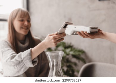 Blonde attractive woman paying with cellphone resting in cafe while waitress holding payment terminal. Girl relaxing in restaurant. Focus on part payment terminal and mobile phone.
