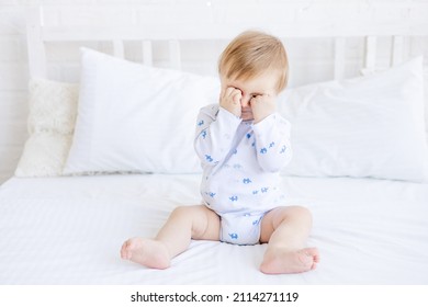 a blonde baby boy rubs sore inflamed eyes or wants to sleep on a white crib while sitting in a room at home with white cotton bedding, the concept of children's goods and accessories - Powered by Shutterstock