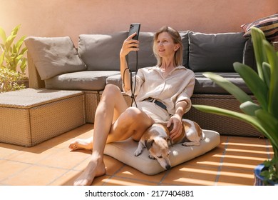 Blonde Beautiful Slim Woman Having Video Call At Summer Sunny Terrace With Senior Dog Jack Russell Terrier. Using Cell Phone For Online Dating. Long Legs. Beige Clothes 