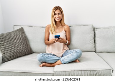 Blonde beautiful young woman sitting on the sofa at home using smartphone with a happy and cool smile on face. lucky person. 