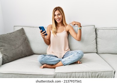 Blonde beautiful young woman sitting on the sofa at home using smartphone looking confident with smile on face, pointing oneself with fingers proud and happy. 