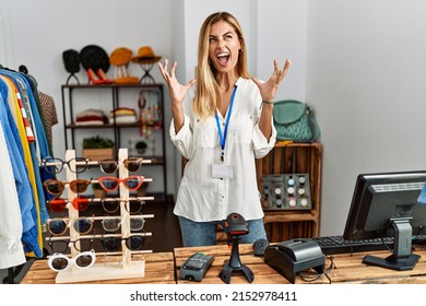 Blonde beautiful young woman working as manager at retail boutique crazy and mad shouting and yelling with aggressive expression and arms raised. frustration concept. 