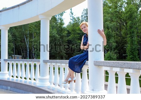 Blonde in a blue dress sits on a white balustrade in a park on a summer day