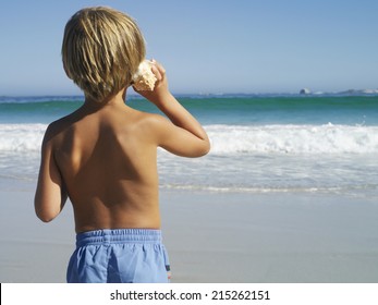 Blonde boy (5-7) standing on beach, listening to sea shell, looking at horizon over sea, rear view