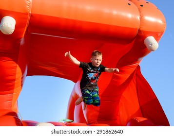 Blonde Boy (7-9 Years) Wearing Black T-shirt And Multi-colored Camouflage Shorts Jumping In Air On Red Bouncy Castle, Blue Sky In Background