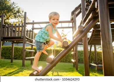 A Blonde Boy Smiles While Climbing Up The Ladder Of A Wooden Jungle Gym In A Play Park In The Golden Afternoon Sun.