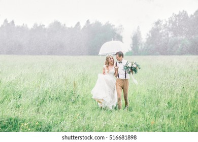 The Blonde Bride And Groom Are On The Field In The Rain. Wedding On A Rainy Day.