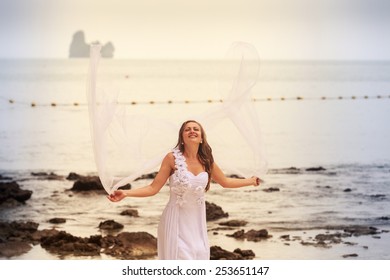 blonde bride in wedding dress jump with white veil stand barefoot on a sand beach against cliff and island sea water background - Powered by Shutterstock