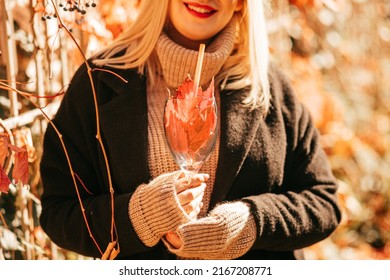 Blonde With Bright Red Lipstick On Her Lips In Black Coat Holds Glass With Autumn Cocktail Of Bright Orange Leaves Against Background Of Yellow-orange Bushes. Colors Of Autumn. Seasons.