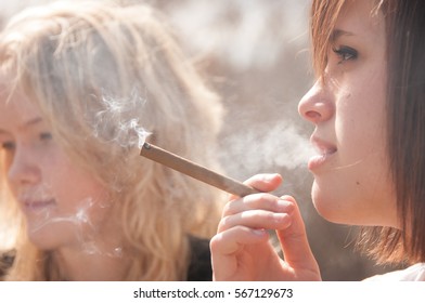 Blonde And Brunette Girls Smoking Thin Cigars Outside. Focus On Brunette Girl Int He Foreground.  Blonde Girl In The Background Out Of Focus, Shallow Depth Of Field.  Cute Girl Smokes A Cigar Outside.