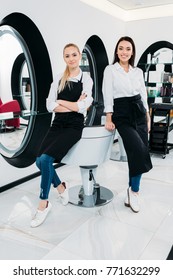 Blonde And Brunette Hairdressers Leaning On Hairdresser Chair At Salon