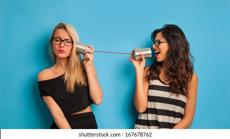 Blonde and brunette women talking with tin can telephone against blue background. 