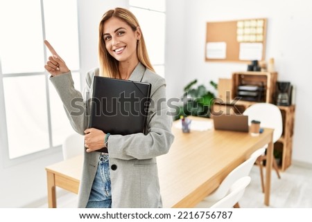 Blonde business woman at the office cheerful with a smile on face pointing with hand and finger up to the side with happy and natural expression 