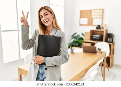 Blonde business woman at the office smiling looking to the camera showing fingers doing victory sign. number two. 