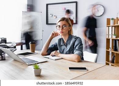 blonde businesswoman in eyeglasses sitting at workplace with gadgets in open space office, motion blur - Shutterstock ID 1856495932