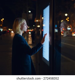 Blonde kaukasische Geschäftsfrau berühren sensiblen Bildschirm der intelligenten City-Bushaltestelle in der Nachtstadt mit Licht außerhalb des Fokus, junge Frau, die Internet-Geld-Zahlung mit automatisiertem Teller-Rechner