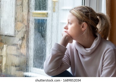 Blonde Caucasian female seated indoors next to a wet window day dreaming.  - Powered by Shutterstock