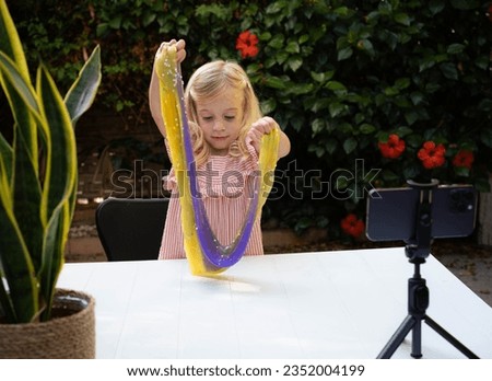 Blonde caucasian girl playing with slime and watching video lesson on smart phone. Online education, classes outdoors in garden, on distance. Child sensory development.