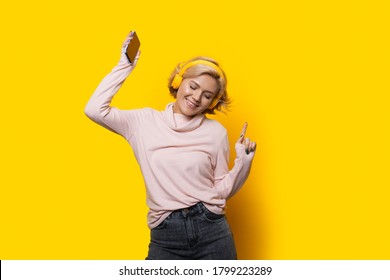 Blonde caucasian woman with headphones is dancing on a yellow background smiling and holding a phone