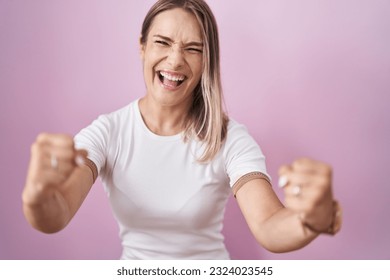 Blonde caucasian woman standing over pink background angry and mad raising fists frustrated and furious while shouting with anger. rage and aggressive concept.  - Shutterstock ID 2324023545