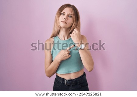 Blonde caucasian woman standing over pink background in hurry pointing to watch time, impatience, looking at the camera with relaxed expression 