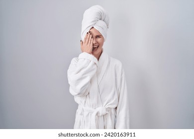 Blonde caucasian woman wearing bathrobe covering one eye with hand, confident smile on face and surprise emotion.  - Powered by Shutterstock