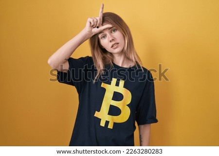 Blonde caucasian woman wearing bitcoin t shirt making fun of people with fingers on forehead doing loser gesture mocking and insulting. 