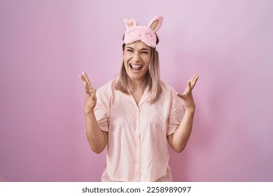 Blonde caucasian woman wearing sleep mask and pajama crazy and mad shouting and yelling with aggressive expression and arms raised. frustration concept.  - Shutterstock ID 2258989097