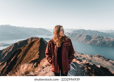 blonde caucasian young woman in profile with hands in pockets of brown jacket black t-shirt and black sweater looking off from a mountain summit calm and relaxed enjoying the surroundings, roys peak - Powered by Shutterstock