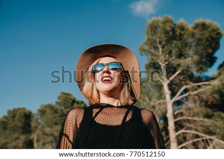 Porträt einer blonden Frau mit Hut, Sonnenbrille und roten Lippen, die bei Sonnenuntergang die Natur genießt.