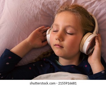 Blonde Child Girl In Headphones Listening To Music For Better Sleep Relaxing Calming Podcast For Stress Relief With Smartphone Online In Bed At Home. Audio Book Tales Against Insomnia For Tired Kids.