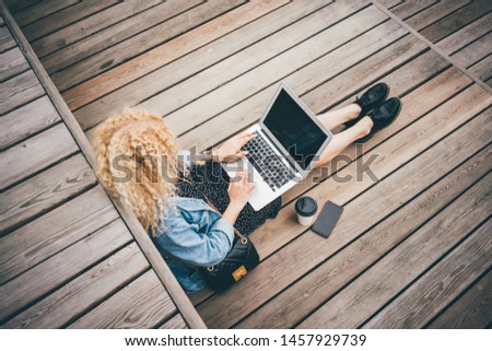 Blonde curly young casually dressed girl using mobile phone and lap top in the park, modern gadget with wifi in public places. Freelancer girl working in the park.