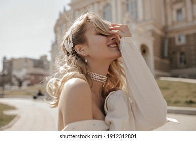 Blonde curly young woman smiles sincerely outdoors. Happy charming girl in white blouse and pearl necklace poses in city center.