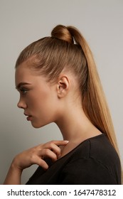 A Blonde European Girl With Long Straight Hair In A High Ponytail And A Hair Knot Is Posing On The Gray Background. The Lady Is Wearing A Black Tee-shirt. 