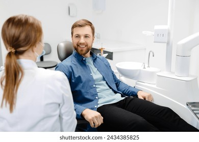 Blonde female dentist in dental office talking with male patient and preparing for treatment. Handsome bearded man in dentist chair looking at his doctor with smile. - Powered by Shutterstock