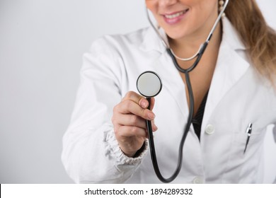 Blonde female doctor with eyeglasses and hair gathered in white coat makes a nice smile while holding stethoscope isolated on white background