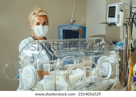 A blonde female doctor in a protective mask caring to a sick baby opening or clothing the incubator. Neonatal intensive care.
