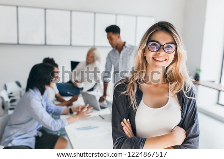 Blonde female executive posing with smile and arms crossed during brainstorm with managers. Indoor portrait of european student spending time in hall with asian and african friends.