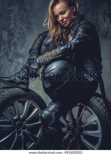 Blonde female in leather clothes sits on a car\'s\
wheel and holds safety\
helmet.