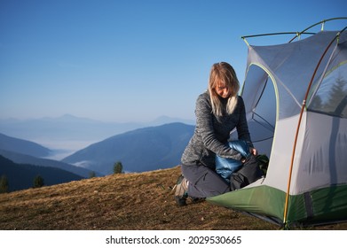 Blonde Female Traveler Packing Her Sleeping Bag In Protective Bag After Night's Rest, Sitting Down At The Edge Of Tent. Vacation In The Mountains In Tourist Campsite. Concept Of Travelling And Camping
