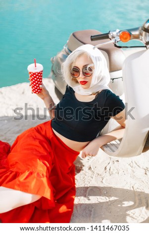 blonde girl 60s in round glasses, white headscarf, black top, long red skirt, white stockings on a sandy beach with blue water on a gray moped under the bright sun summer smile  happy