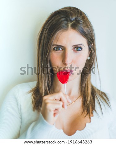 blonde girl biting a red lollipop in the shape of a heart. she has green eyes and is on a white background