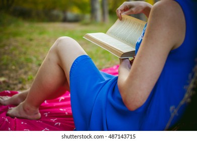 blonde girl in a blue dress sitting on a red blanket on top of the green grass, reading a book in the middle of pine forest trees